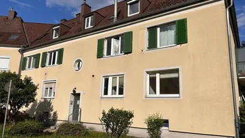 Gelbe Fassade eines Mehrfamilienhauses mit grünen Fensterläden und Satteldach, umgeben von Grünflächen.