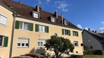 Gelbes Mehrfamilienhaus mit grünen Fensterläden und Satteldach unter blauem Himmel.