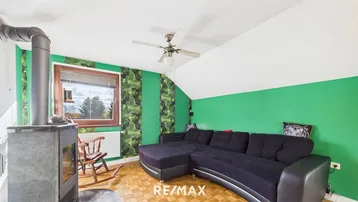Gemütliches Wohnzimmer mit schwarzem Sofa, grün gestrichenen Wänden und einem Holzofen unter der Dachschräge.