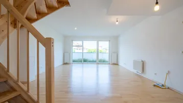 Helles, leeres Wohnzimmer mit Holztreppe, Parkettboden und Zugang zu einem Balkon oder einer Terrasse.