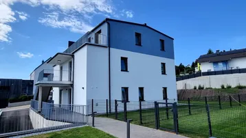 Modernes Mehrfamilienhaus mit blauer und weißer Fassade, umgeben von Grünflächen und einem Gehweg.