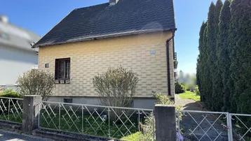 Gelbes Einfamilienhaus mit Satteldach und kleinen Fenstern, umgeben von Hecken und einem Zaun.