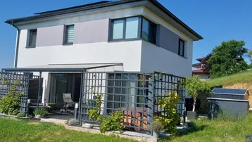 Modernes Haus mit überdachter Terrasse, Whirlpool und grüner Umgebung unter blauem Himmel.
