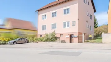 Mehrfamilienhaus mit hellrosa Fassade und Parkplätzen vor dem Gebäude unter blauem Himmel.