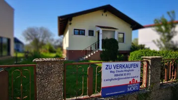 Einfamilienhaus mit gelber Fassade und einem 'Zu Verkaufen'-Schild im Vorgrund.
