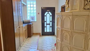 Traditioneller Flur mit Kachelofen, Holzschränken und gemusterten Fliesen, der zum Eingangsbereich führt.