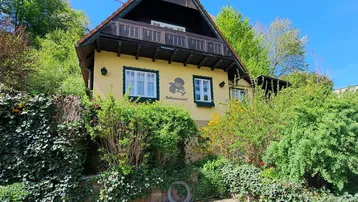Traditionelles Haus mit gelber Fassade, Holzbalkon und üppiger grüner Umgebung am Hang.