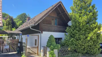 Rustikales Holzhaus mit Satteldach und grüner Umgebung unter blauem Himmel.