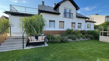 Weißes Einfamilienhaus mit gepflegtem Garten, Terrasse und Balkon unter blauem Himmel.