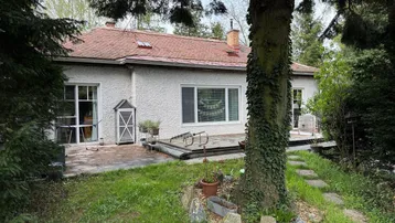 Rückseite des Hauses mit einer gepflasterten Terrasse, umgeben von einem grünen Garten und Bäumen.
