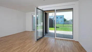 Helles Zimmer mit Holzboden und großen Glastüren, die zum Garten und einer kleinen Terrasse führen.