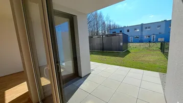 Helle Terrasse mit Fliesenboden und Blick auf den Garten sowie angrenzende Gebäude.