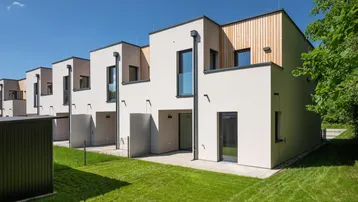 Moderne Reihenhäuser mit weißer Fassade, Holzverkleidung und kleinen Gärten unter blauem Himmel.