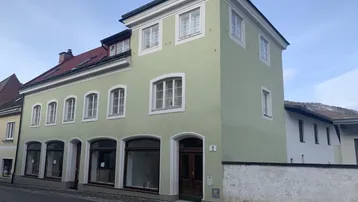 Historisches Gebäude mit hellgrüner Fassade und großen Schaufenstern an einer Straße gelegen.