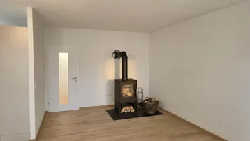 Geräumiges Zimmer mit Holzboden und digital hinzugefügtem Holzofen, bietet gemütliche Atmosphäre.