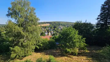Weitläufiger Blick über eine grüne Landschaft mit einem Dorf in der Ferne unter blauem Himmel.