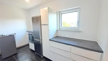 Funktionale Küche mit weißen Unterschränken, dunkler Arbeitsplatte, Kühlschrank und Fenster.