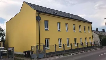 Gelbe Fassade eines Mehrfamilienhauses mit Satteldach und weißen Fenstern an einer ruhigen Straße.
