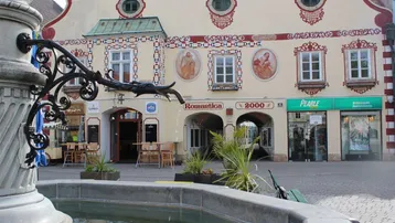 Historisches Gebäude mit reich verzierter Fassade und Geschäften im Erdgeschoss, davor ein Brunnen auf einem Stadtplatz.