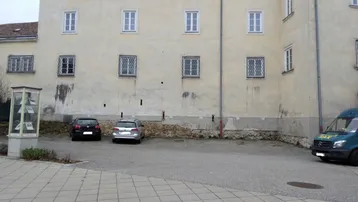 Historische Gebäudefassade mit traditionellen Fenstern und einem gepflasterten Parkplatz davor.