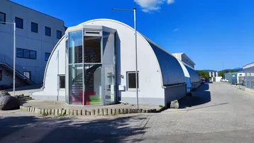 Moderne, bogenförmige Gewerbeimmobilie mit markantem Glaseingang und sichtbarer Wendeltreppe unter blauem Himmel.