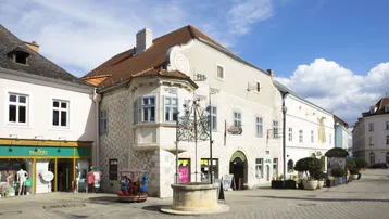 Historisches Gebäude an einem Stadtplatz mit Geschäften und einem Brunnen unter blauem Himmel.