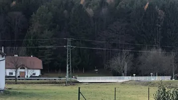 Blick auf ein traditionelles Haus mit rotem Dach, umgeben von grünen Feldern und einem dichten Wald im Hintergrund.