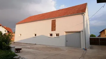 Gebäudeaußenansicht mit rotem Ziegeldach, großer Garagentür und gepflastertem Vorplatz unter bewölktem Himmel.