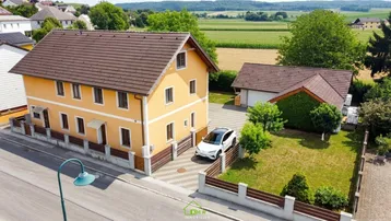 Haus mit gelber Fassade, braunem Dach und gepflasterter Einfahrt, umgeben von Grünflächen.