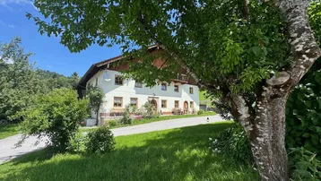 Traditionelles Bauernhaus mit weißer Fassade und Holzbalkonen, umgeben von grüner Natur und Bäumen.