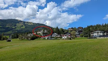 Panoramablick auf ein Dorf in den Bergen, umgeben von grünen Wiesen und Wäldern unter blauem Himmel.