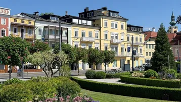 Gelbes Gebäude mit Balkonen und grüner Parkanlage im Vordergrund unter blauem Himmel.