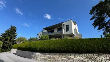 Zeitgenössisches Gebäude mit klaren Linien, umgeben von grüner Vegetation und Hecken unter blauem Himmel.