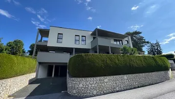 Modernes Mehrfamilienhaus mit gepflegter Hecke und Zufahrt zur Tiefgarage unter blauem Himmel.