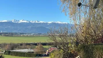 Weitläufiger Blick über grüne Felder und schneebedeckte Berge unter strahlend blauem Himmel.
