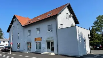 Gebäudeansicht mit hellgrauer Fassade, rotem Ziegeldach und Geschäftsflächen im Erdgeschoss.