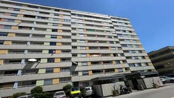 Großes Mehrfamilienhaus mit vielen Balkonen und farbigen Fensterläden unter blauem Himmel.