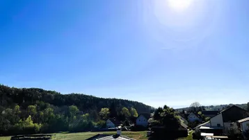 Weitläufiger Panoramablick über grüne Hügel und eine ländliche Wohnsiedlung unter blauem Himmel.