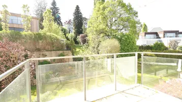 Sonniger Balkon mit Glasgeländer und Blick ins Grüne sowie auf umliegende Gebäude.