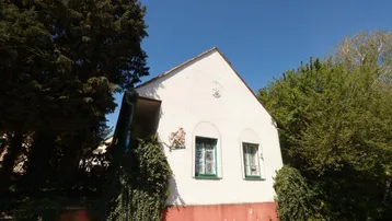 Weiß verputztes Haus mit grünen Fensterrahmen und rotem Sockel in ländlicher Umgebung.
