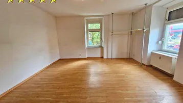 Heller Wohnraum mit Laminatboden, großen Fenstern und einfacher Ausstattung.