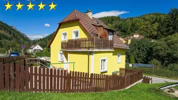 Gelbes Einfamilienhaus mit Holzzaun und Balkon in ländlicher Umgebung mit Bergblick.