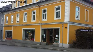 Das Bild zeigt die gelbe Fassade eines Altbaus mit weißen Fenstern und Ladenflächen im Erdgeschoss an einer Straße.