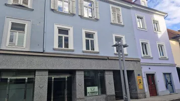 Ansicht der Gebäudefassade mit Geschäften im Erdgeschoss und Wohnungen darüber, mit blauen und lila Farbtönen.