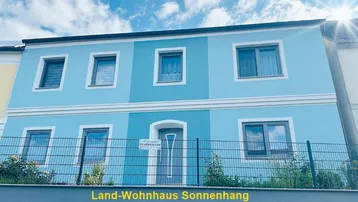Vorderansicht eines Land-Wohnhauses mit hellblauer Fassade und weißen Akzenten unter blauem Himmel.