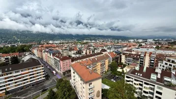 Panoramablick über eine Stadt mit Wohngebäuden und Bergen im Hintergrund unter bewölktem Himmel.