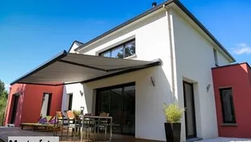 Modernes Haus mit großer Holzterrasse, Markise und Sitzbereich unter blauem Himmel.