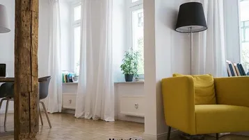 Helles Wohnzimmer mit Holzbalken, gelbem Sessel und großen Fenstern mit weißen Vorhängen.