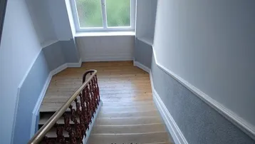 Helles Treppenhaus mit Holztreppe, Fenster und hellgrauen Wänden, das zu einem oberen Stockwerk führt.