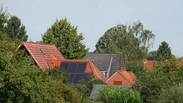 Blick über Dächer von Wohnhäusern, umgeben von üppigem Grün und Bäumen, mit Solaranlagen auf einem Dach.